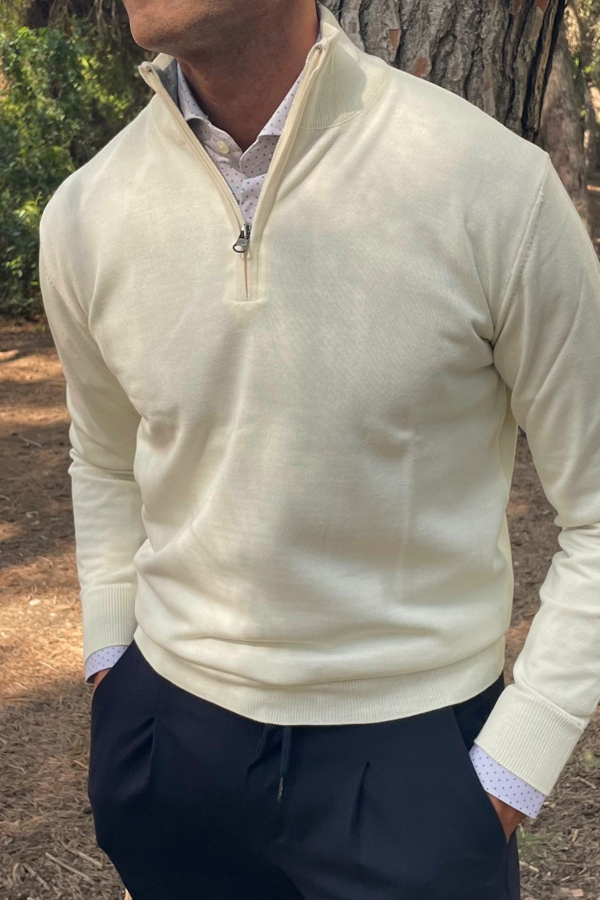 Jersey de hombre blanco nata con cuello alto y cremallera, combinado con camisa estampada y pantalón azul marino.
